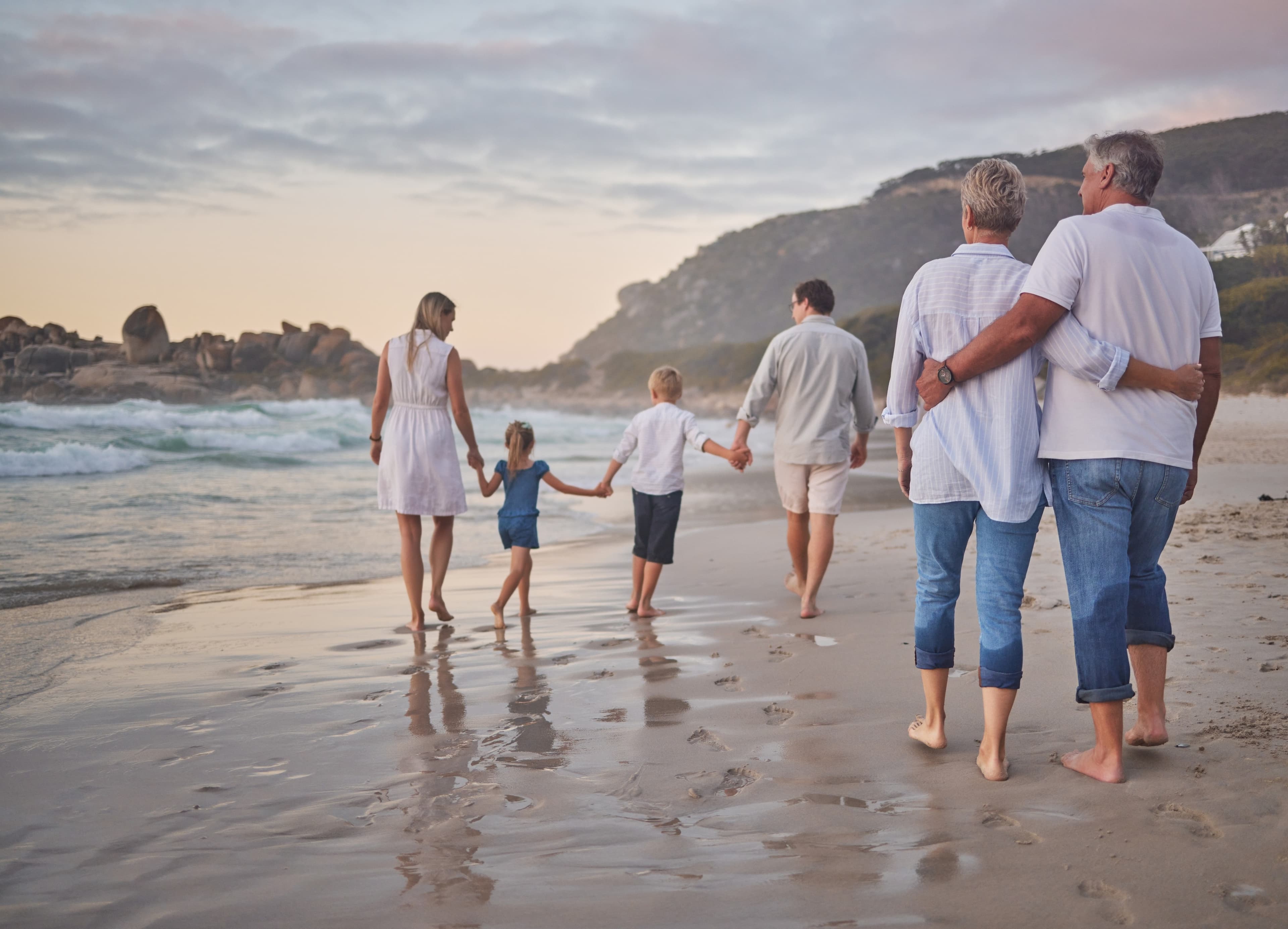 A multi-generational family walks together along the beach at sunset, holding hands and viewed from behind.